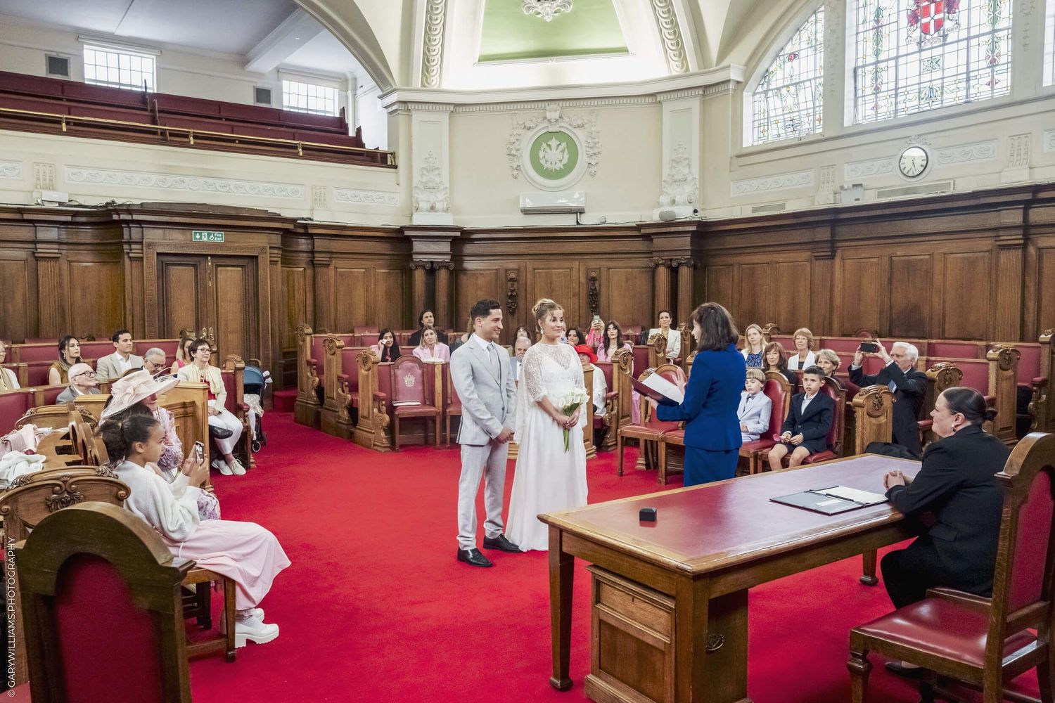 A solemn civil ceremony takes place in an elegant wood-paneled room with red carpeting and stained glass windows.