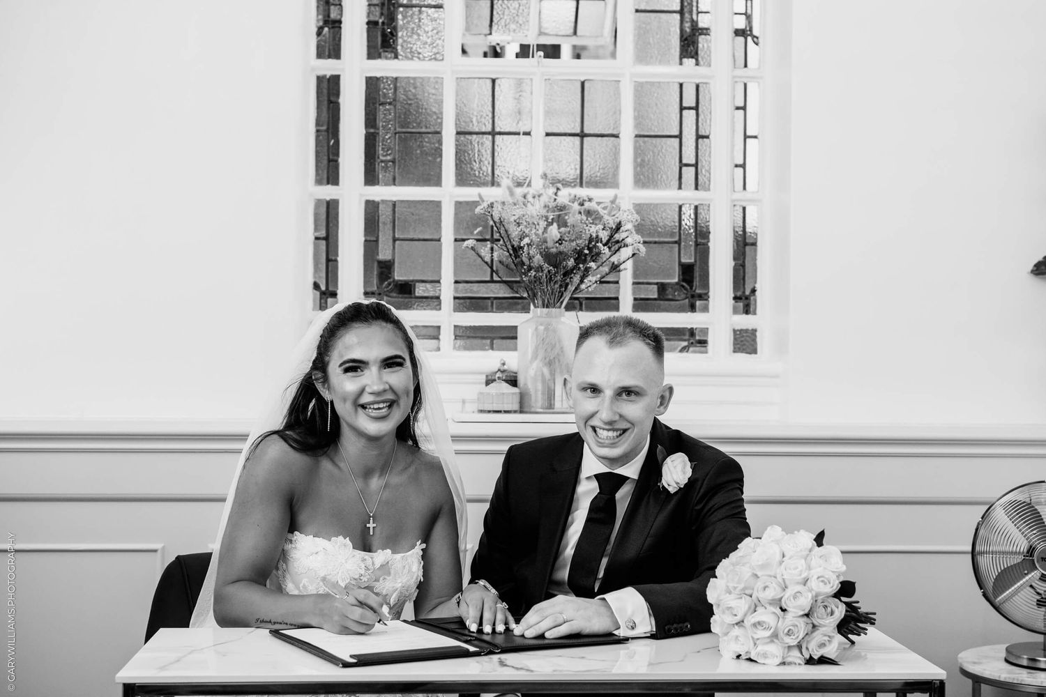 Black and white photo of newlyweds signing marriage documents with joyful expressions.
