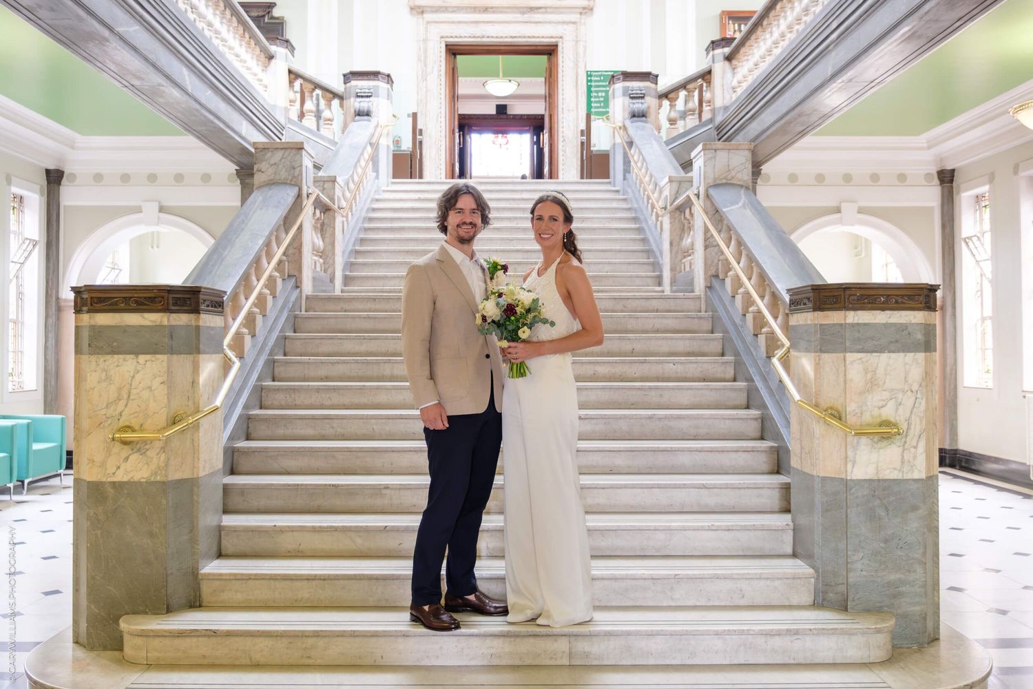 Two wedding guests pose together on a grand marble staircase in an elegant historic building interior.