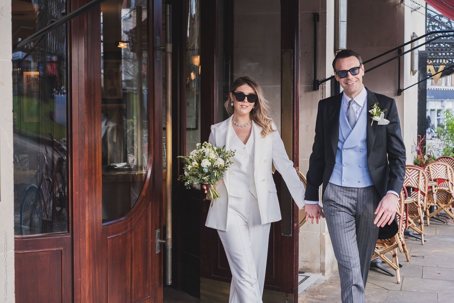 A couple in stylish suits exits a building together, with the person in white holding a floral bouquet.