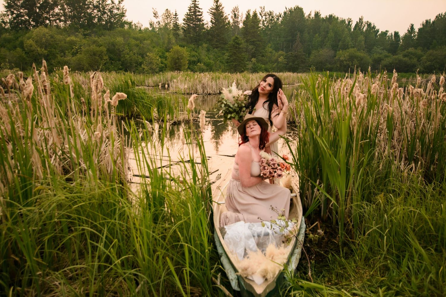 A scenic summer sunset scene at a serene pond surrounded by tall grass and evergreen trees in the background.
