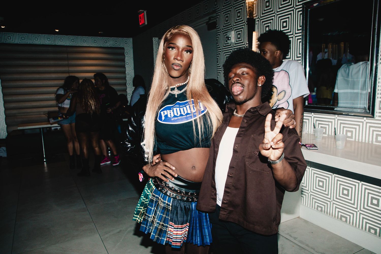 Two people pose together at a nightclub, one wearing a blue crop top and plaid skirt, the other in a brown jacket.