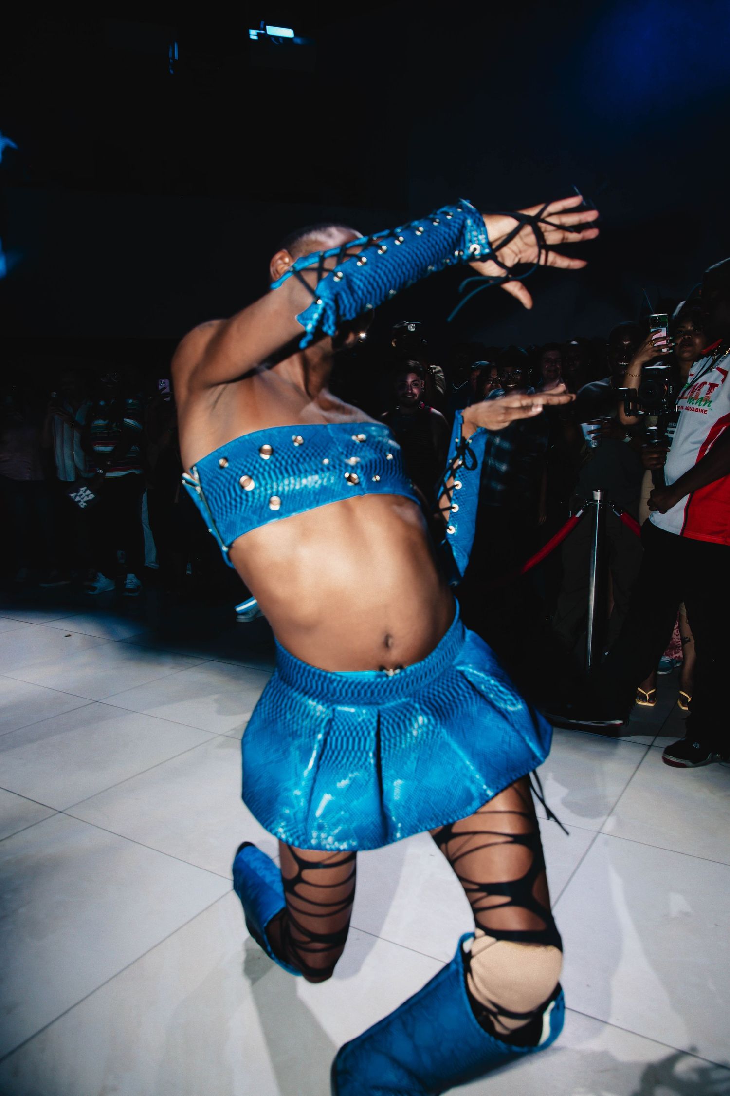 Dancer in sparkly blue outfit performs breakdance moves on white dance floor while crowd watches in background.