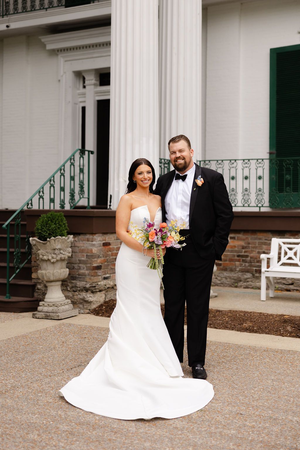 A bride and groom pose together outside a white columned building entrance with a colorful wedding bouquet. Top 10 Nashville Wedding Venues