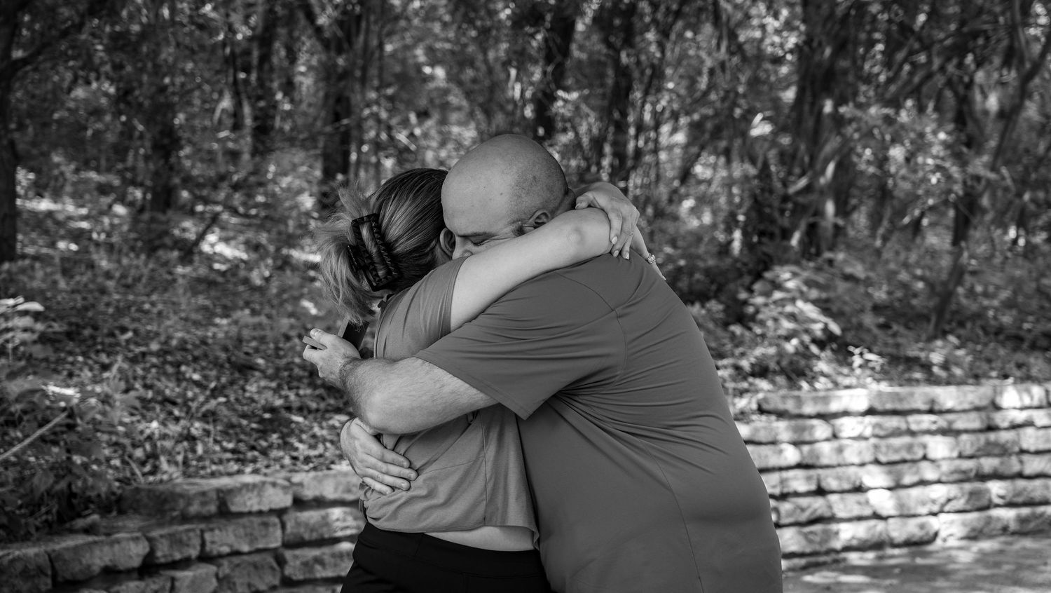 Two figures share an emotional embrace on a brick ledge in a wooded area, captured in black and white photography.