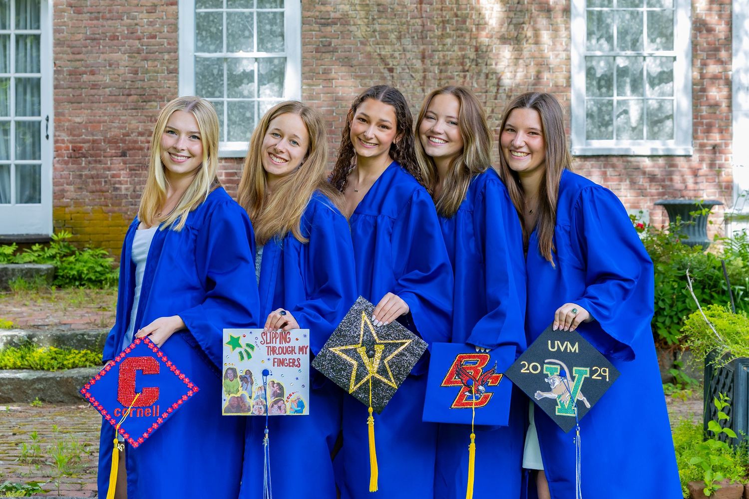 Graduates in bright blue commencement robes display their decorated mortarboard caps outside a brick building.