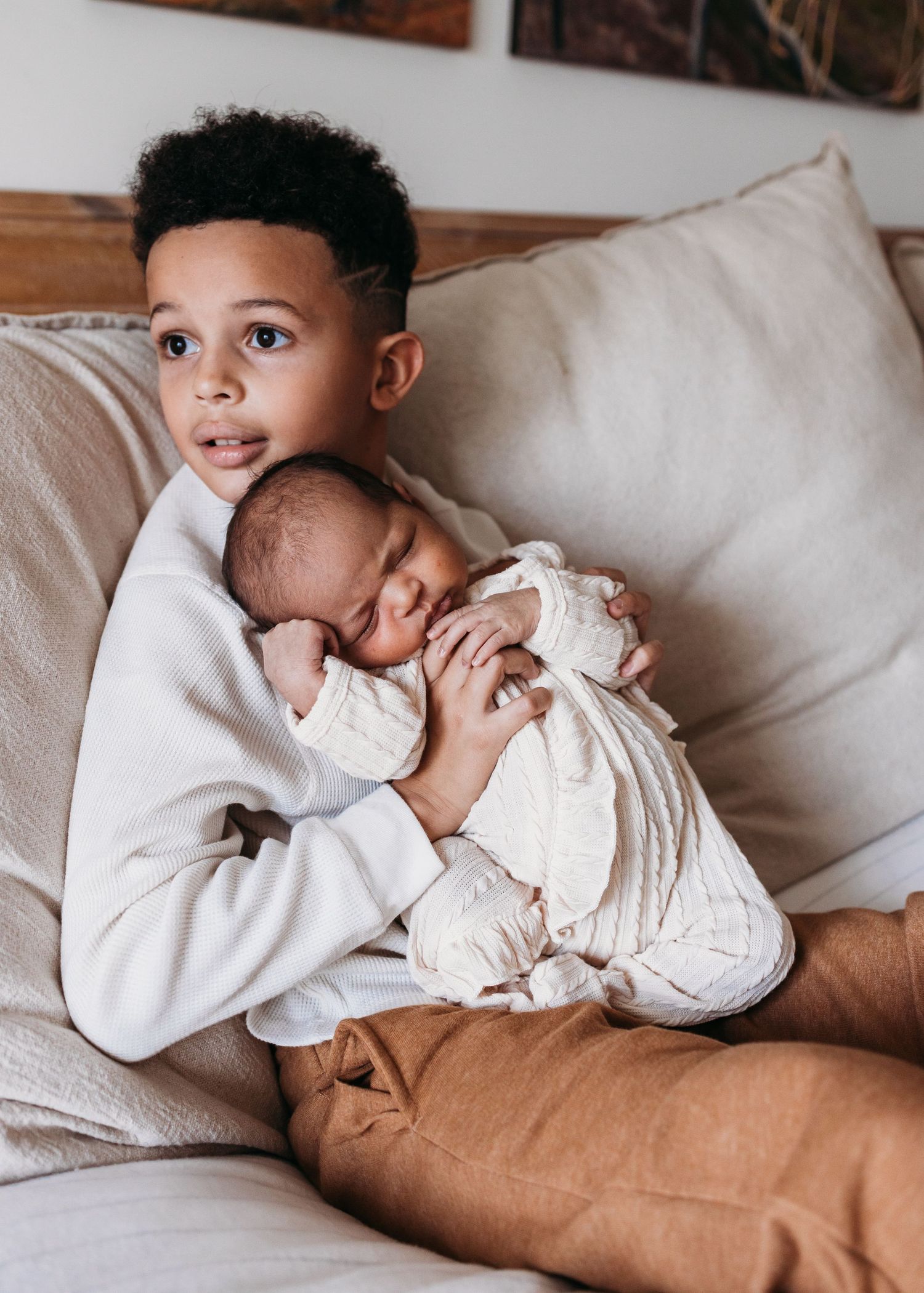 Toddler in white sweatshirt tenderly holds swaddled sleeping newborn while sitting on beige couch.