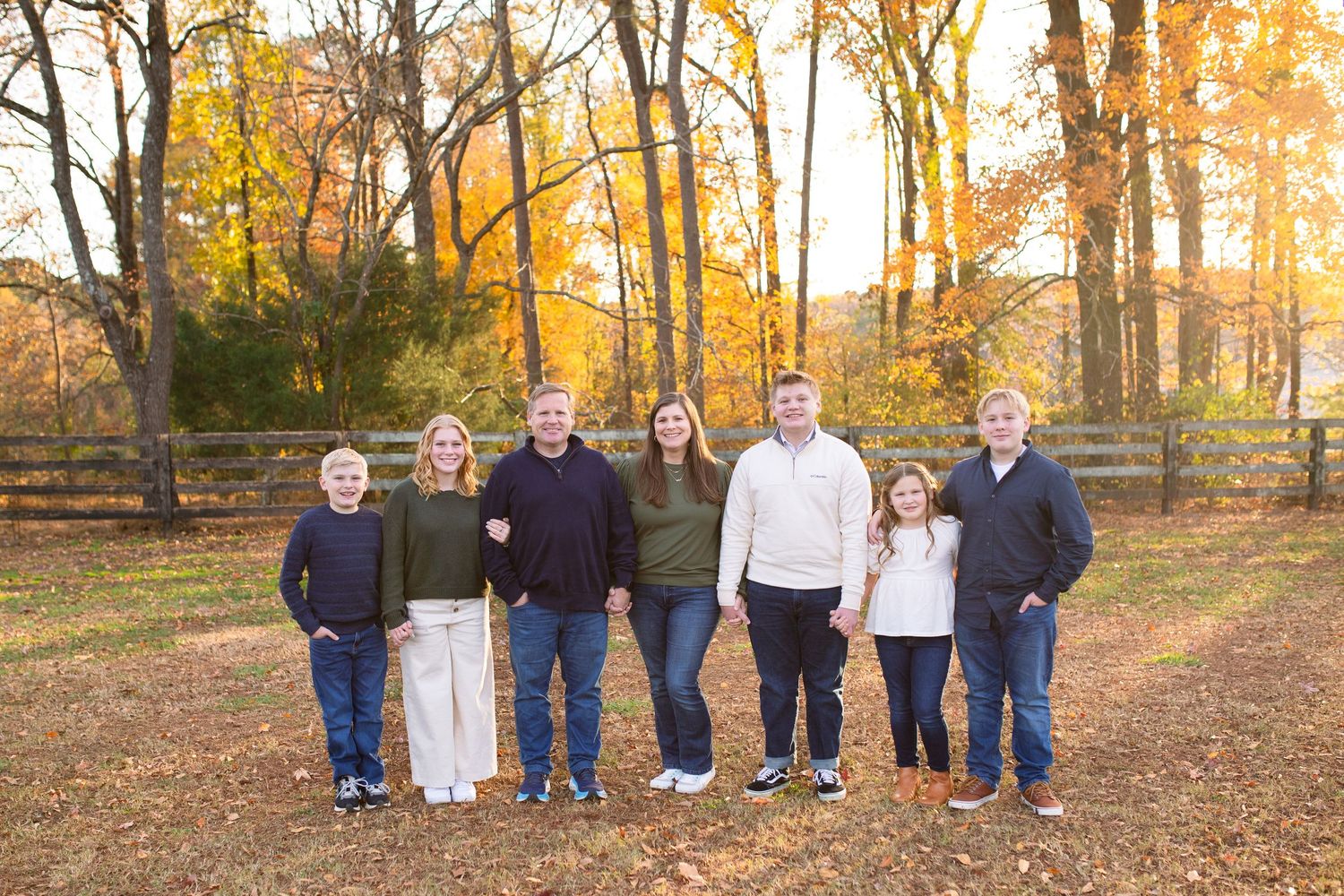 family stands together on fallen autumn leaves with golden trees and sunset light glowing in forest background at Sugg Farm in Holly Springs