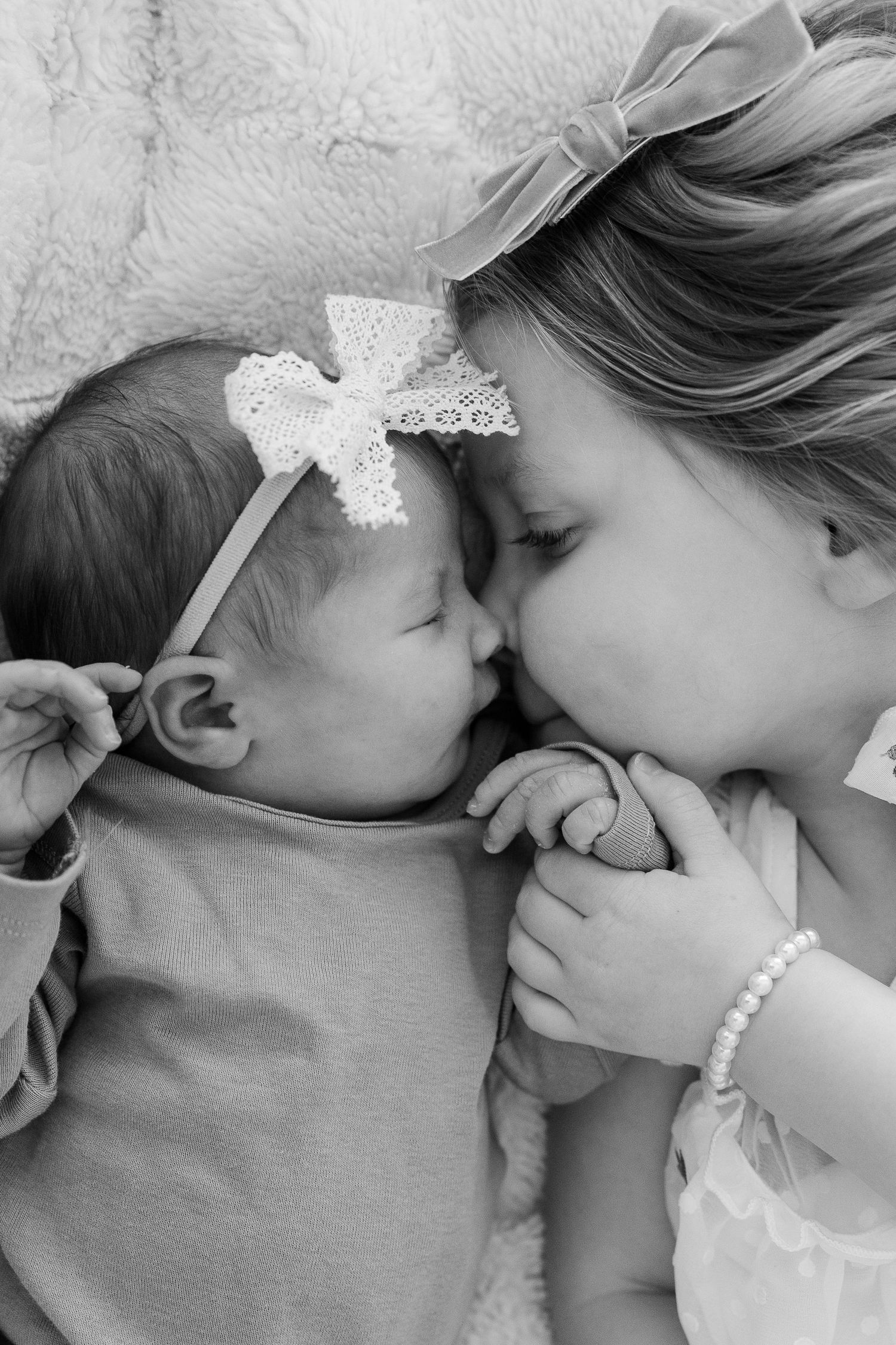 newborn and big sis laying on a blanket nose to nose. both have pretty bows. photo is in black and white.