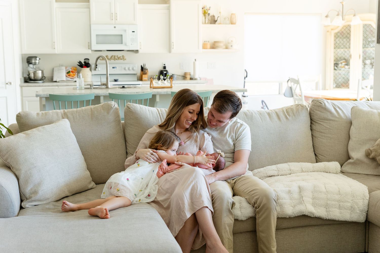 Mom and dad sitting on a cream colored couch holding a newborn in moms arm and a little girl laying in moms other arm toughing newborns head
