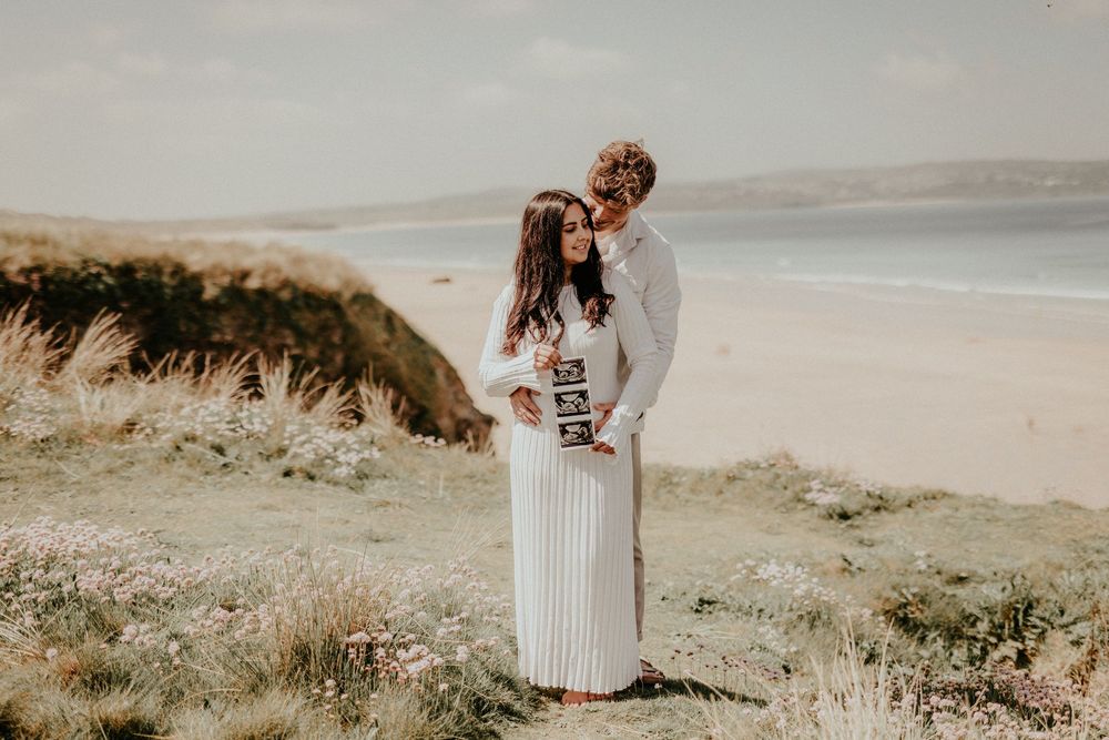 Couple embraces on scenic coastal cliff overlooking sandy beach in dreamy natural lighting.