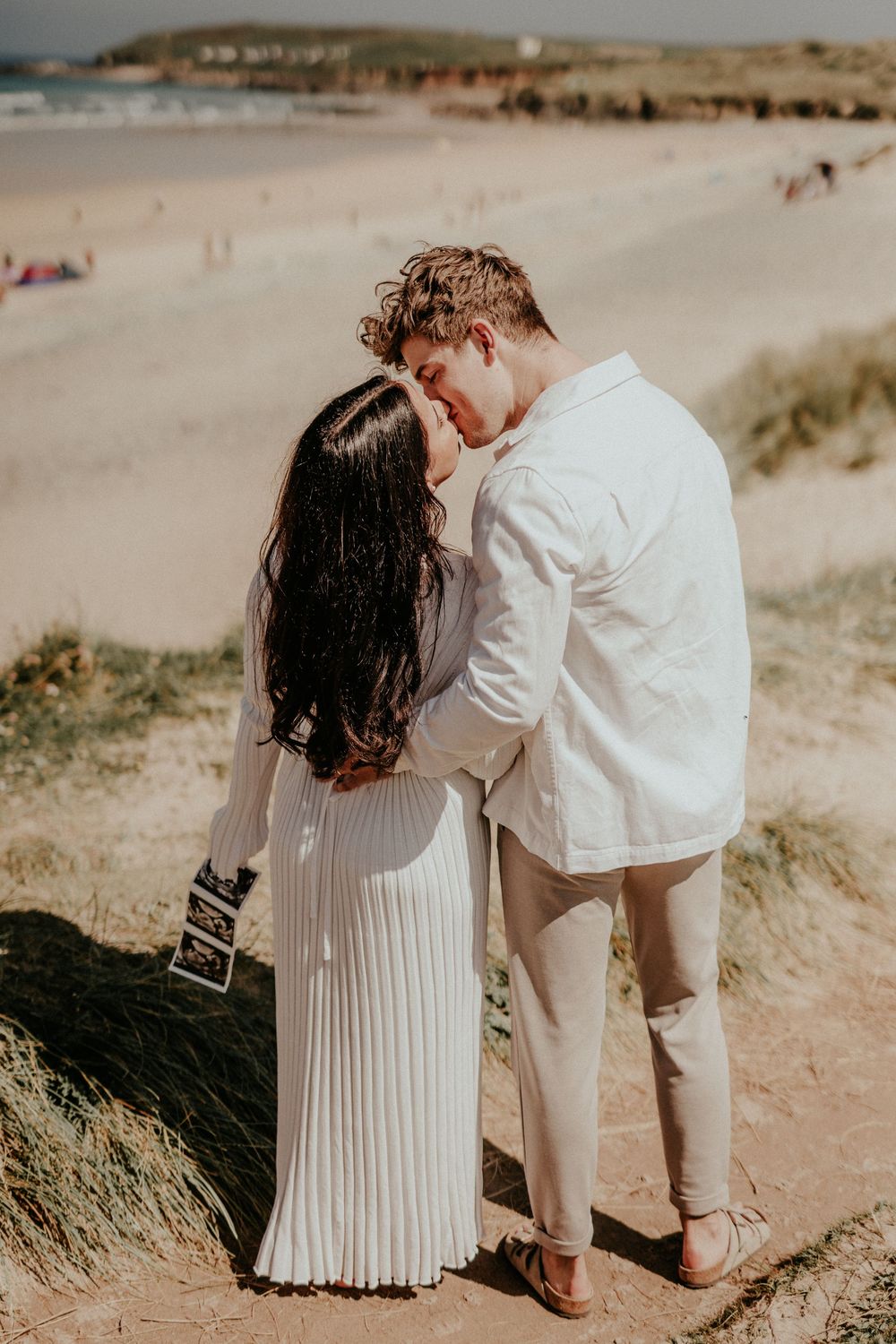 A couple shares an intimate moment on a sandy hill overlooking a beach on a warm sunny day.