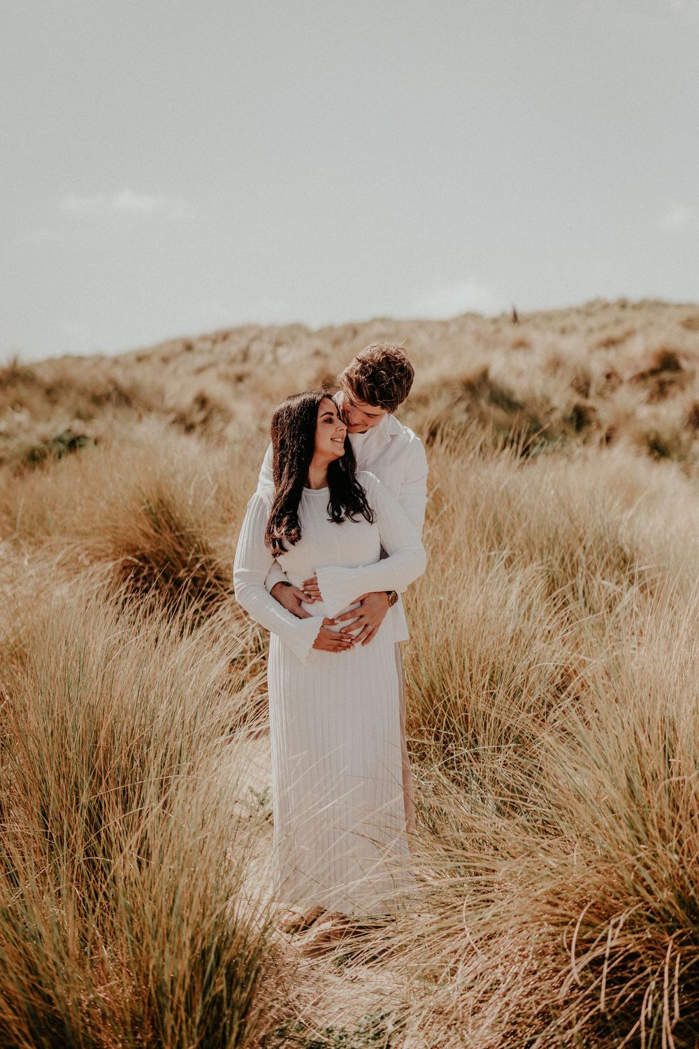 A romantic coastal photography session in warm sepia tones with tall grass swaying in the natural landscape.