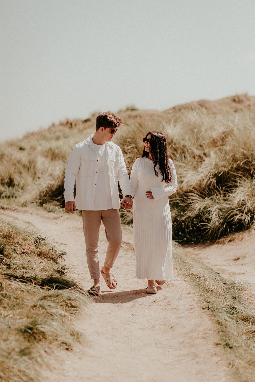A couple in coordinating white outfits walk hand in hand along a sandy nature trail during a warm sunset.