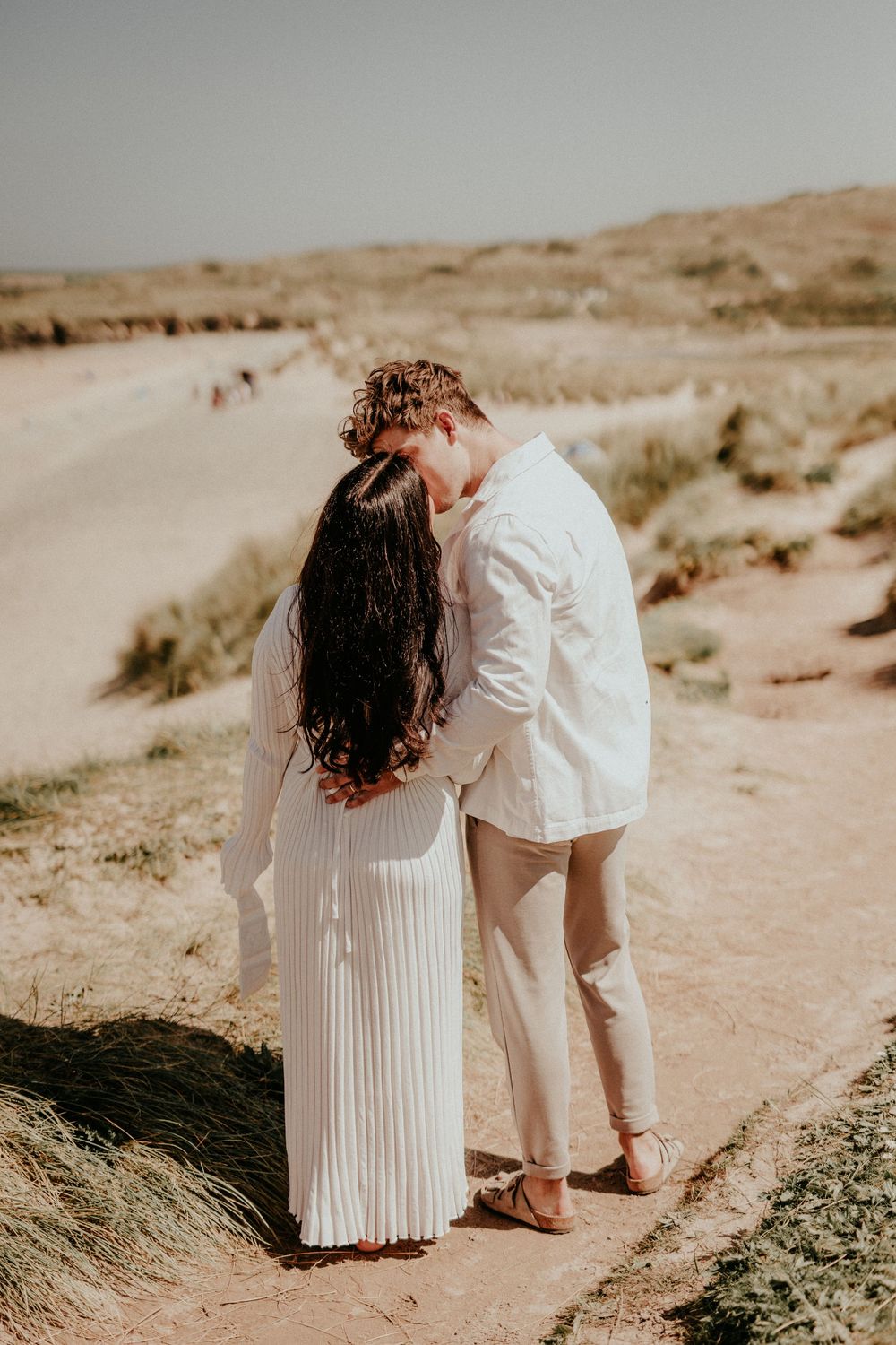 A romantic couple in white outfits walks along a sandy desert landscape with gentle dunes in the background.