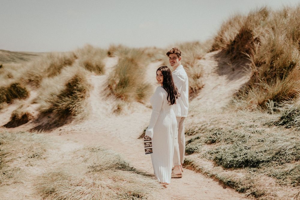 A couple in wedding attire embraces on a sandy path through coastal dunes during a sunset photo session.