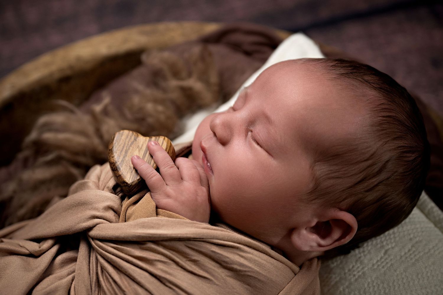 A sleeping newborn baby wrapped in a tan swaddle blanket rests peacefully against a white pillow.
