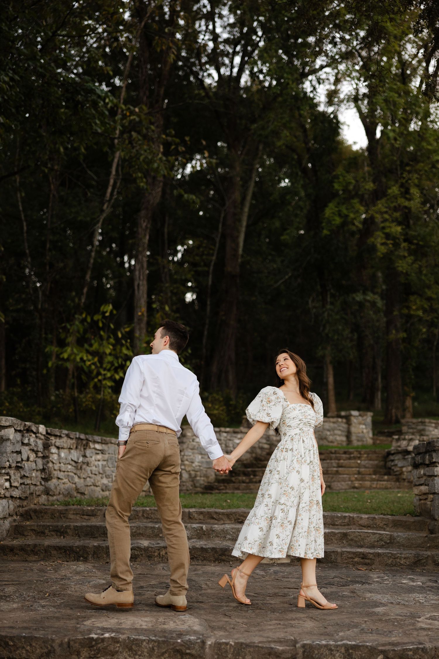 A couple dances playfully together on a stone patio surrounded by trees in a candid engagement photo series.