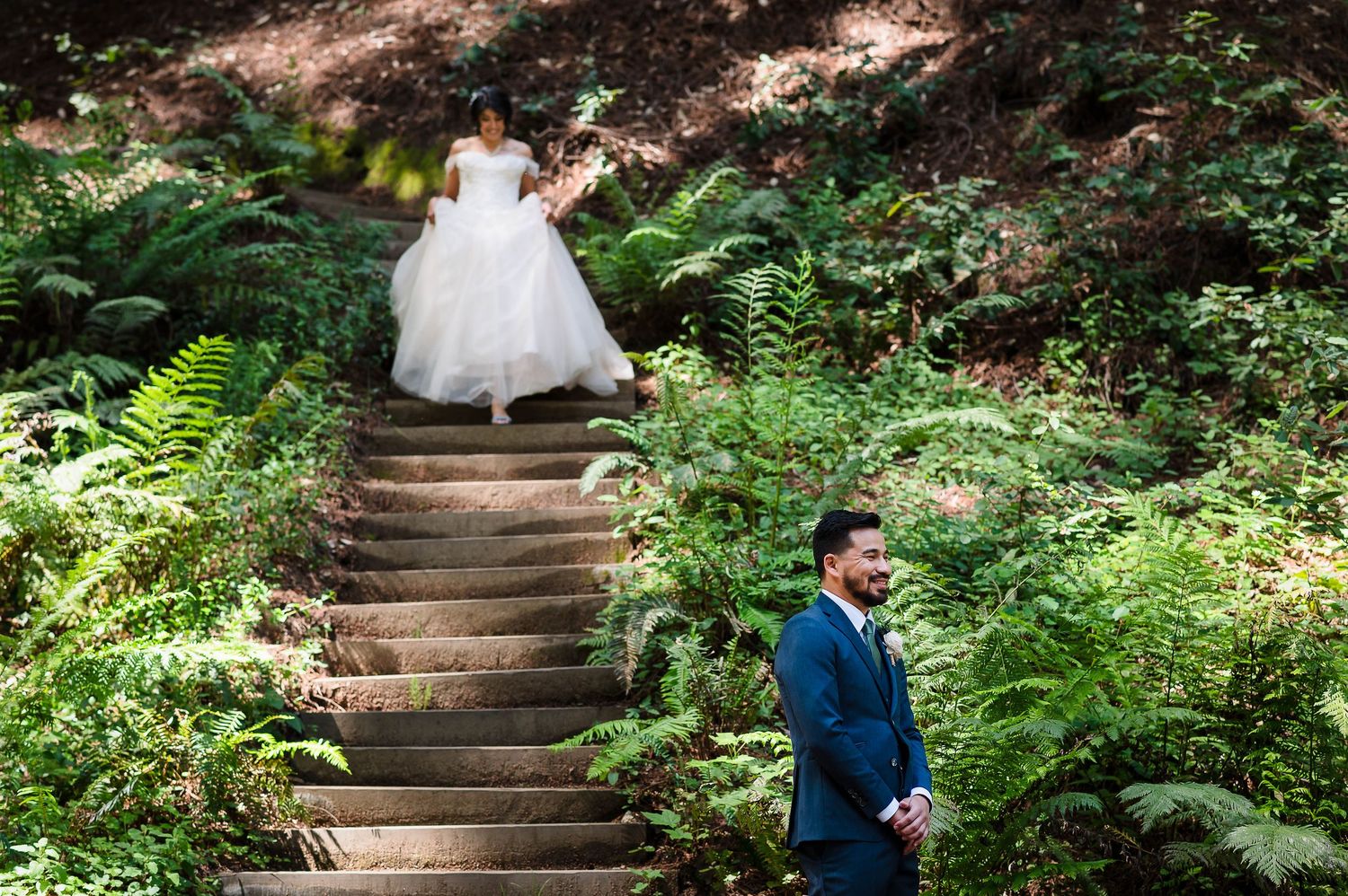Two people in wedding attire stand on stone stairs surrounded by lush ferns in a forest setting.