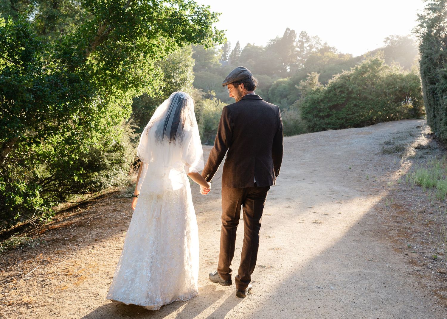 A couple in wedding attire walks hand in hand down a sunlit garden path while wearing bride and groom formalwear.