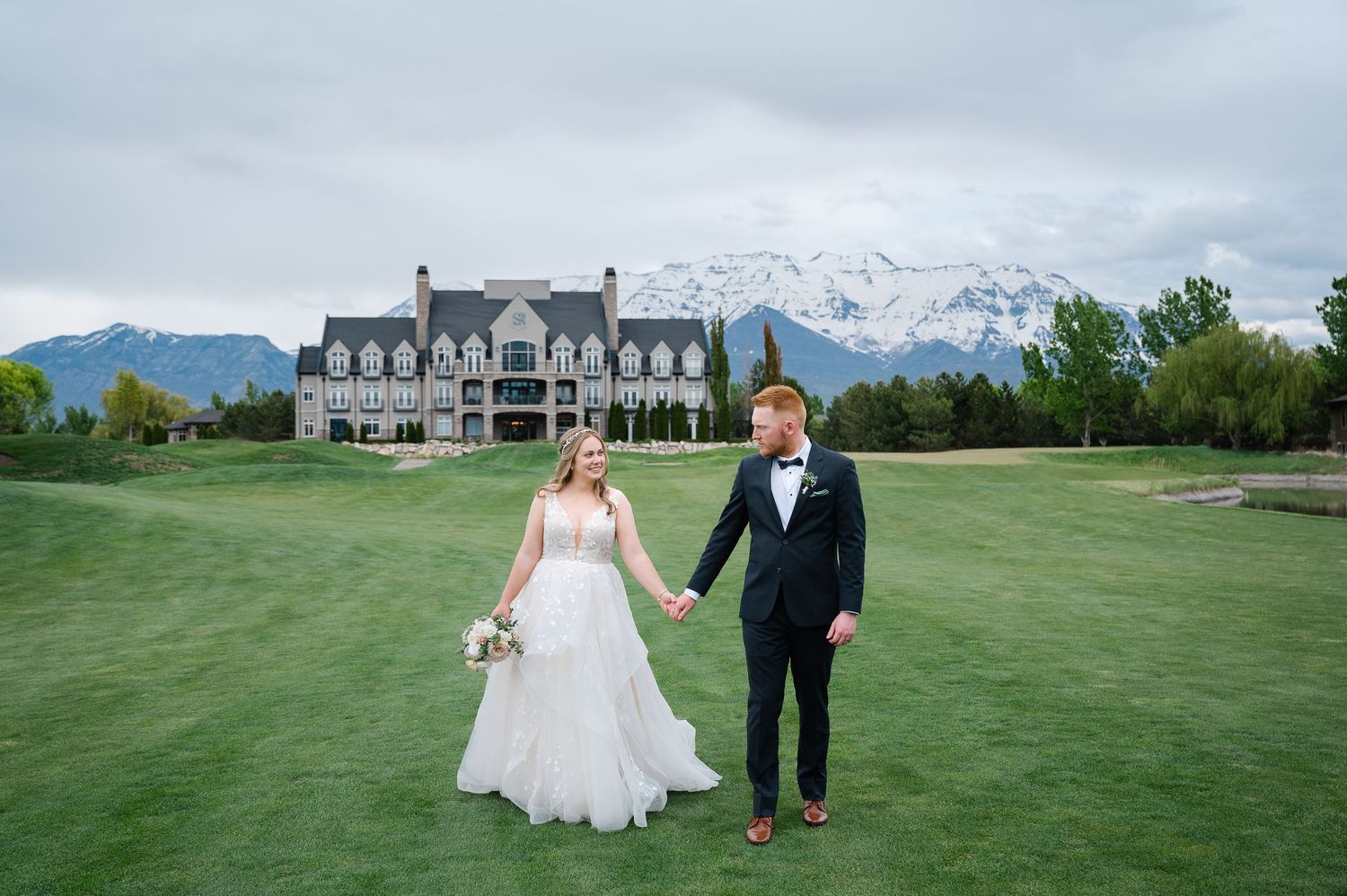 Elegant wedding couple walks on a lush green lawn with a grand mansion and snow-capped mountains in the background.