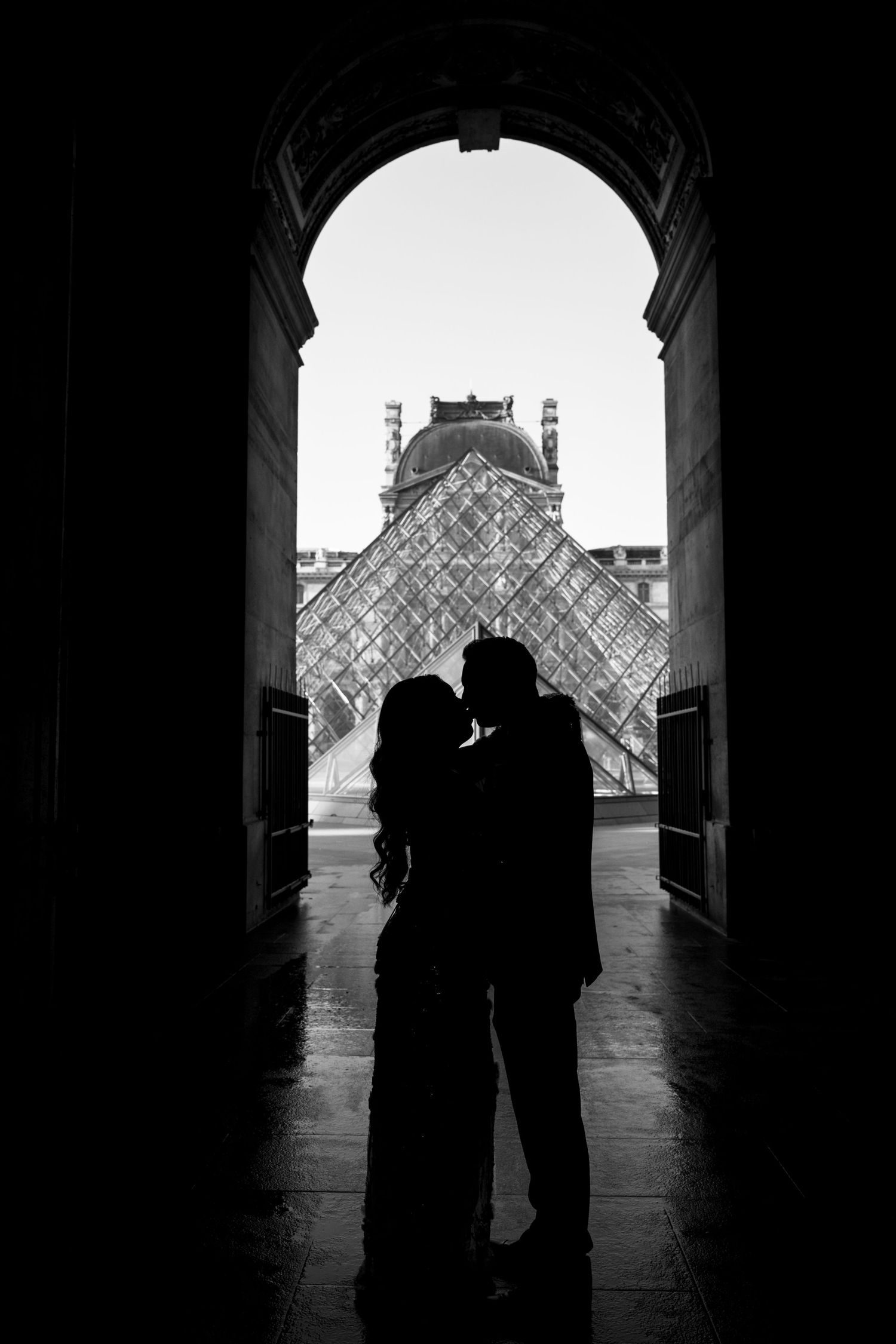 A couple poses under an arched colonnade with palm trees visible in the background at sunset.