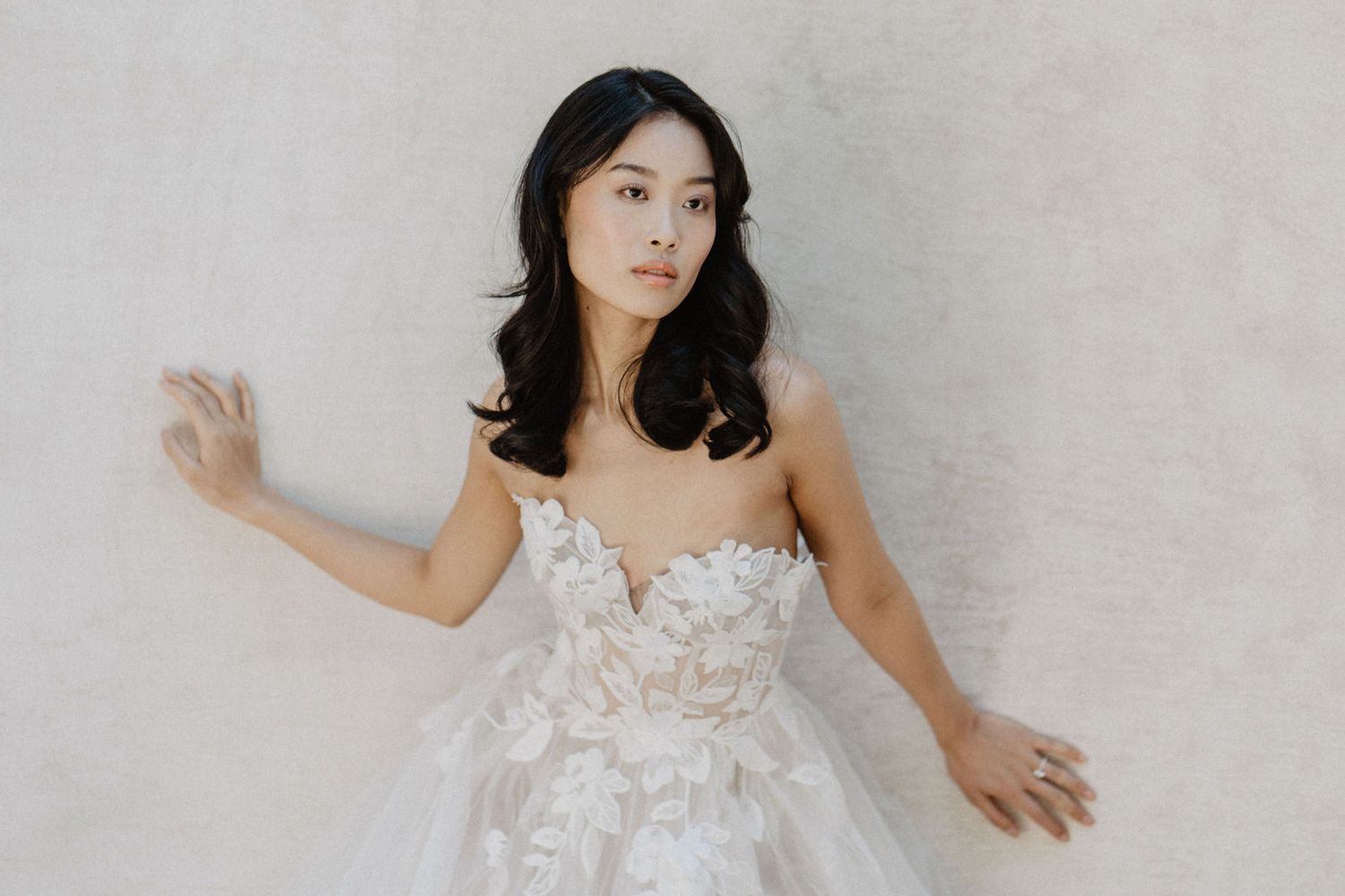 A bridal fashion portrait against a neutral wall showcasing a detailed lace wedding gown.
