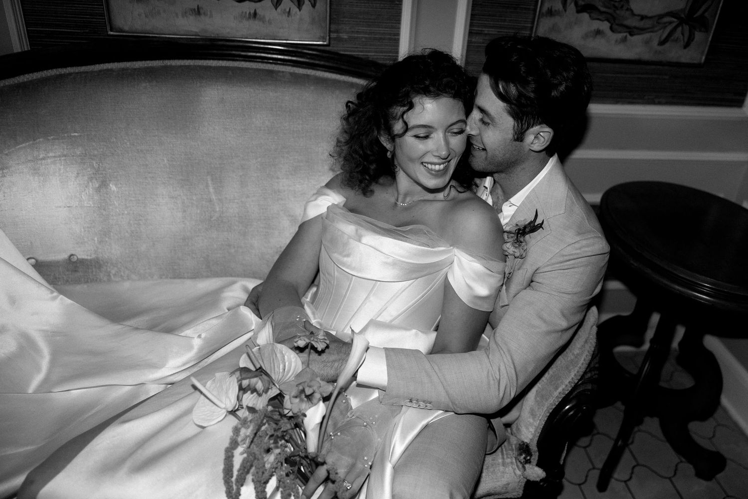 Newlywed couple shares intimate moment on vintage couch with bride holding bouquet in white gown and groom in beige suit.