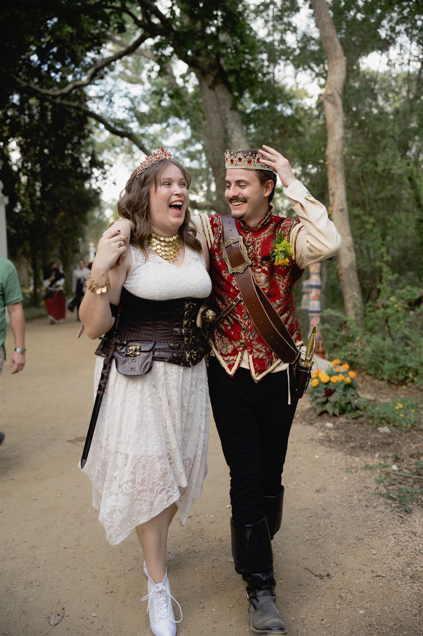 Two joyful people in Renaissance-style costumes share a laugh while walking along a woodland path at a festival.