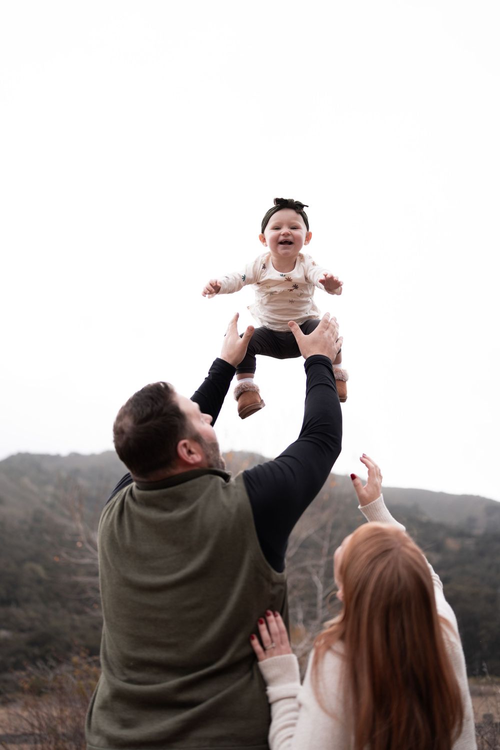 Parents playfully lift baby in the air against mountainous backdrop during outdoor photoshoot.