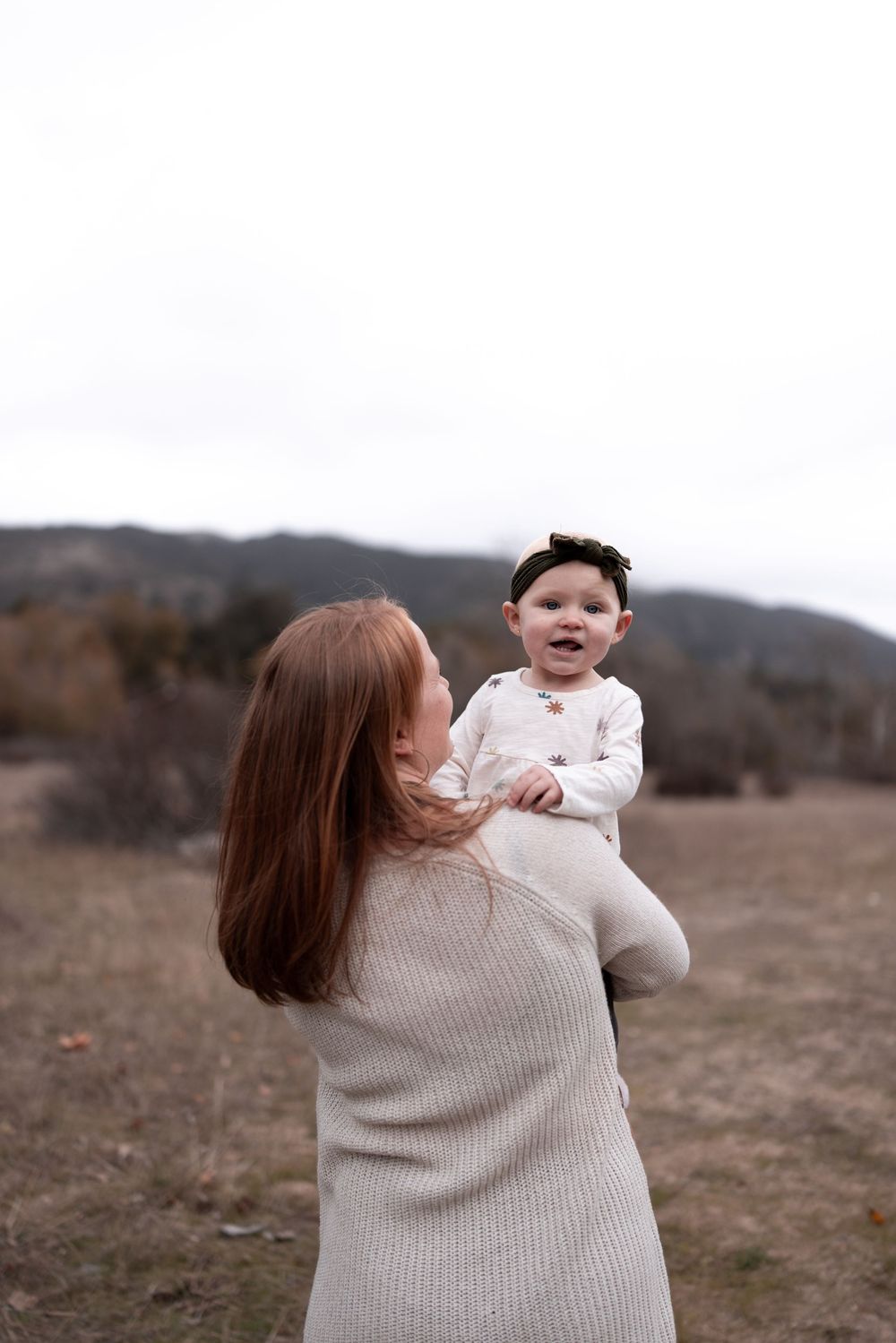 A parent in cream sweater lifts a small child wearing white against mountains and cloudy sky.