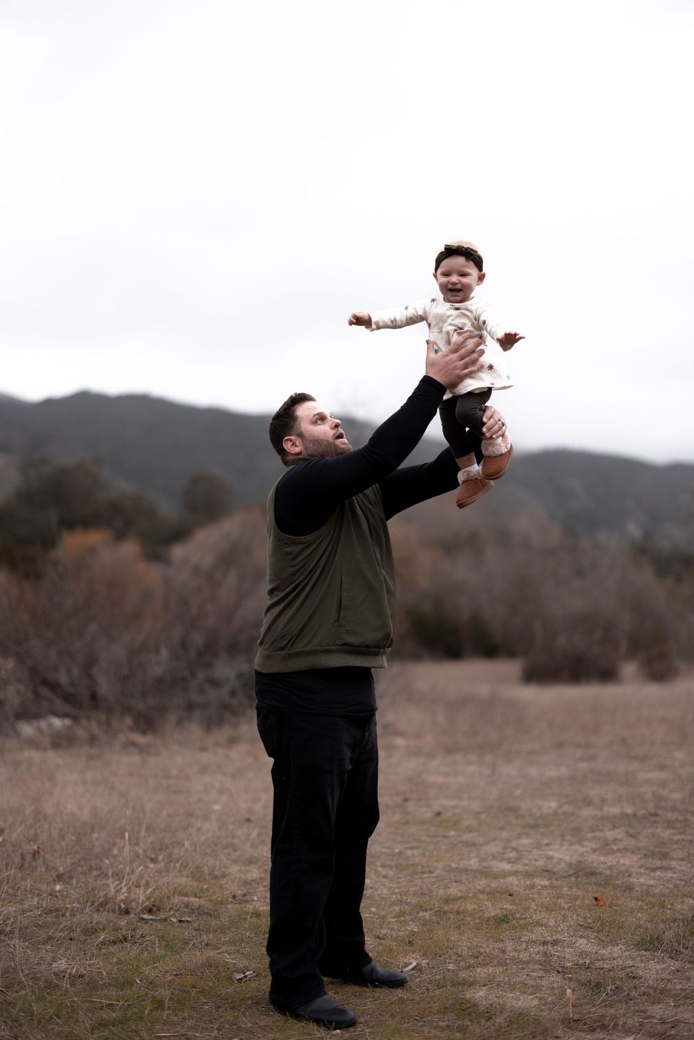 A parent lifts small child high in the air during playful moment outdoors against mountains.