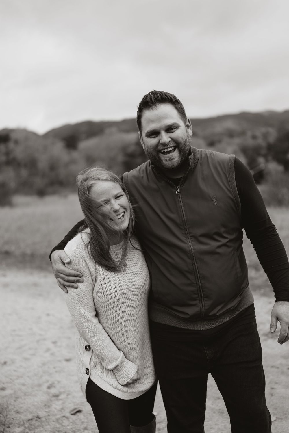 Happy couple embraces and laughs together on a dirt path with rolling hills in the background.