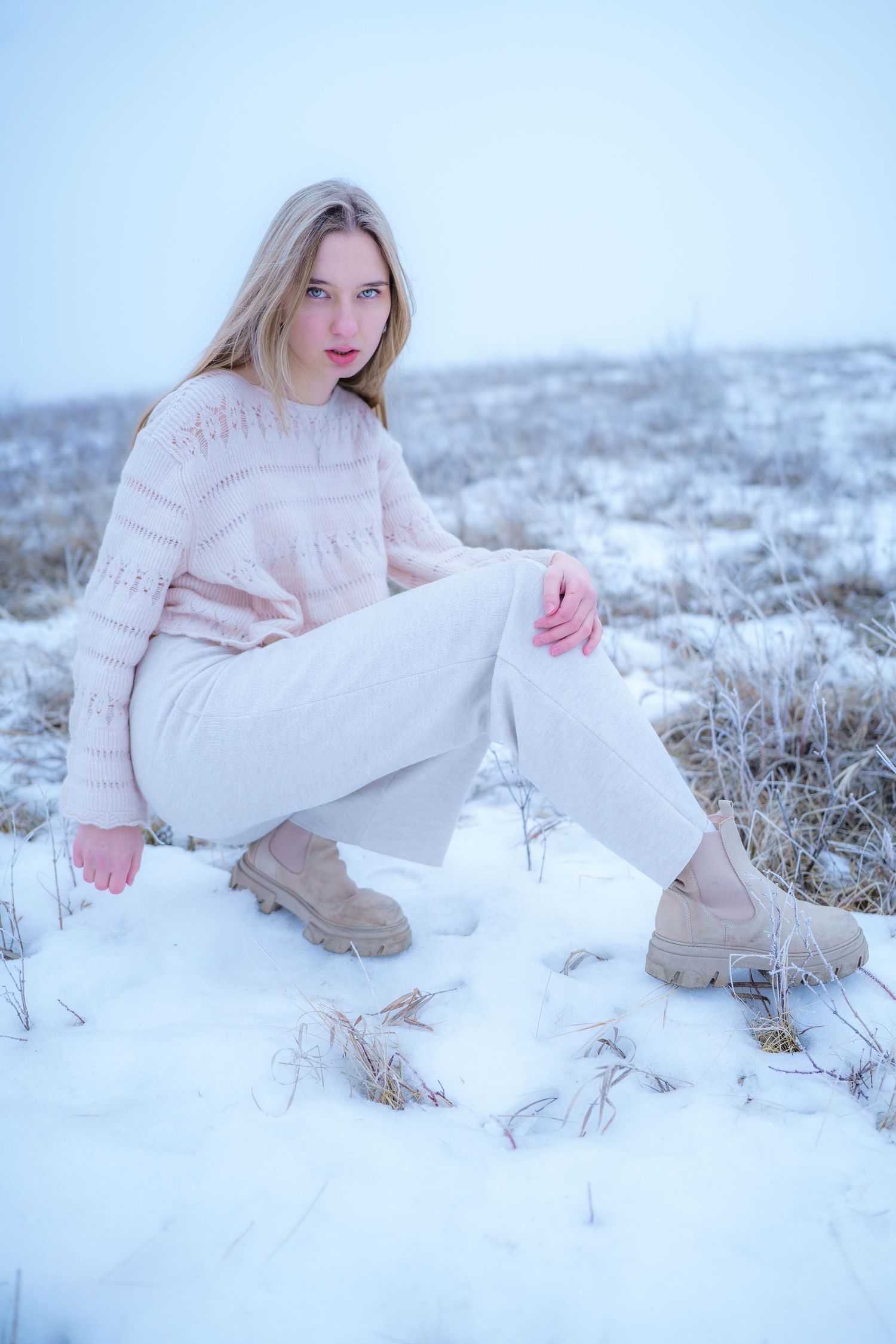 Person in winter fashion wearing cream sweater and white pants poses on snowy ground in muted tones.