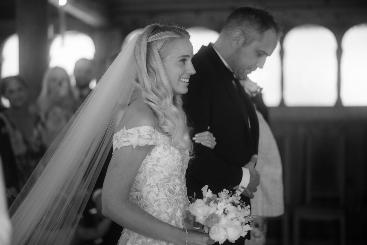 A bride in a beautiful lace dress smiles while holding her bouquet during the wedding ceremony.