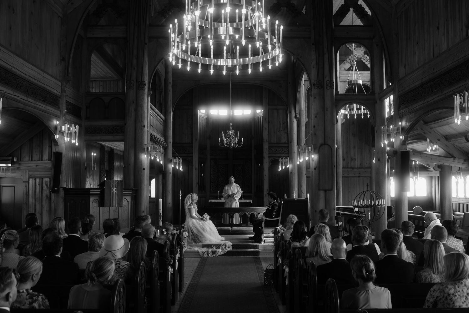 Church wedding ceremony under a grand chandelier with guests seated in wooden pews.