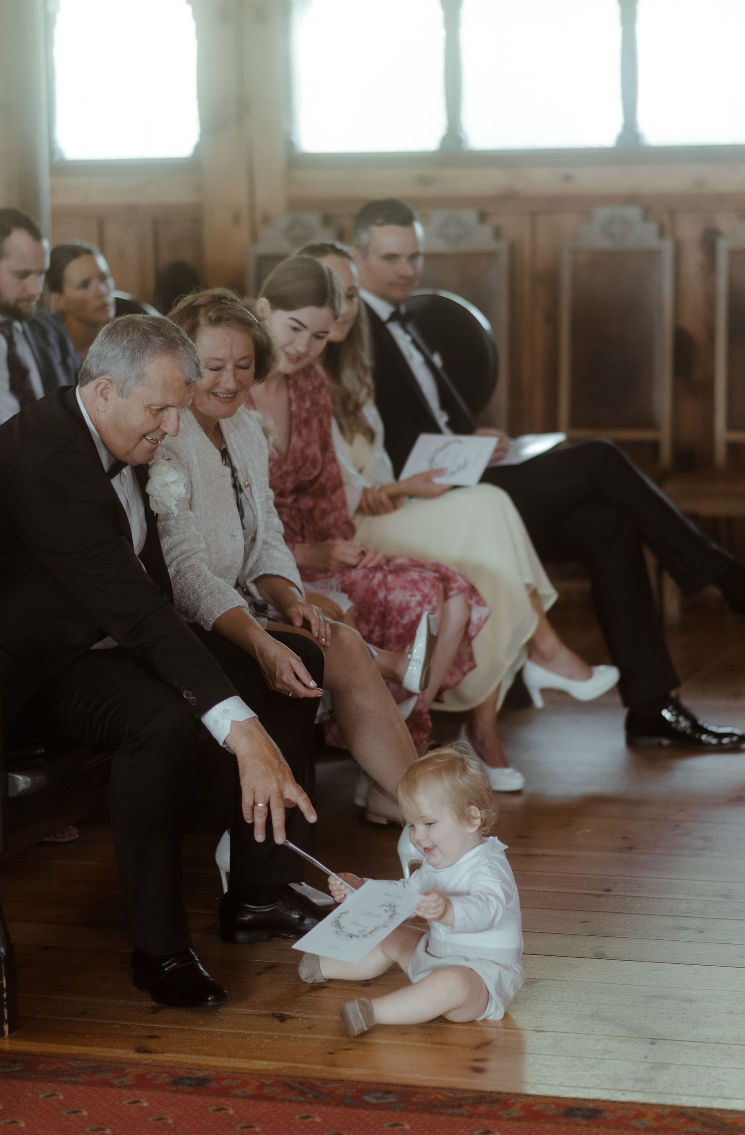 Wedding guests in formal attire watch a small child playing on wooden church floor.