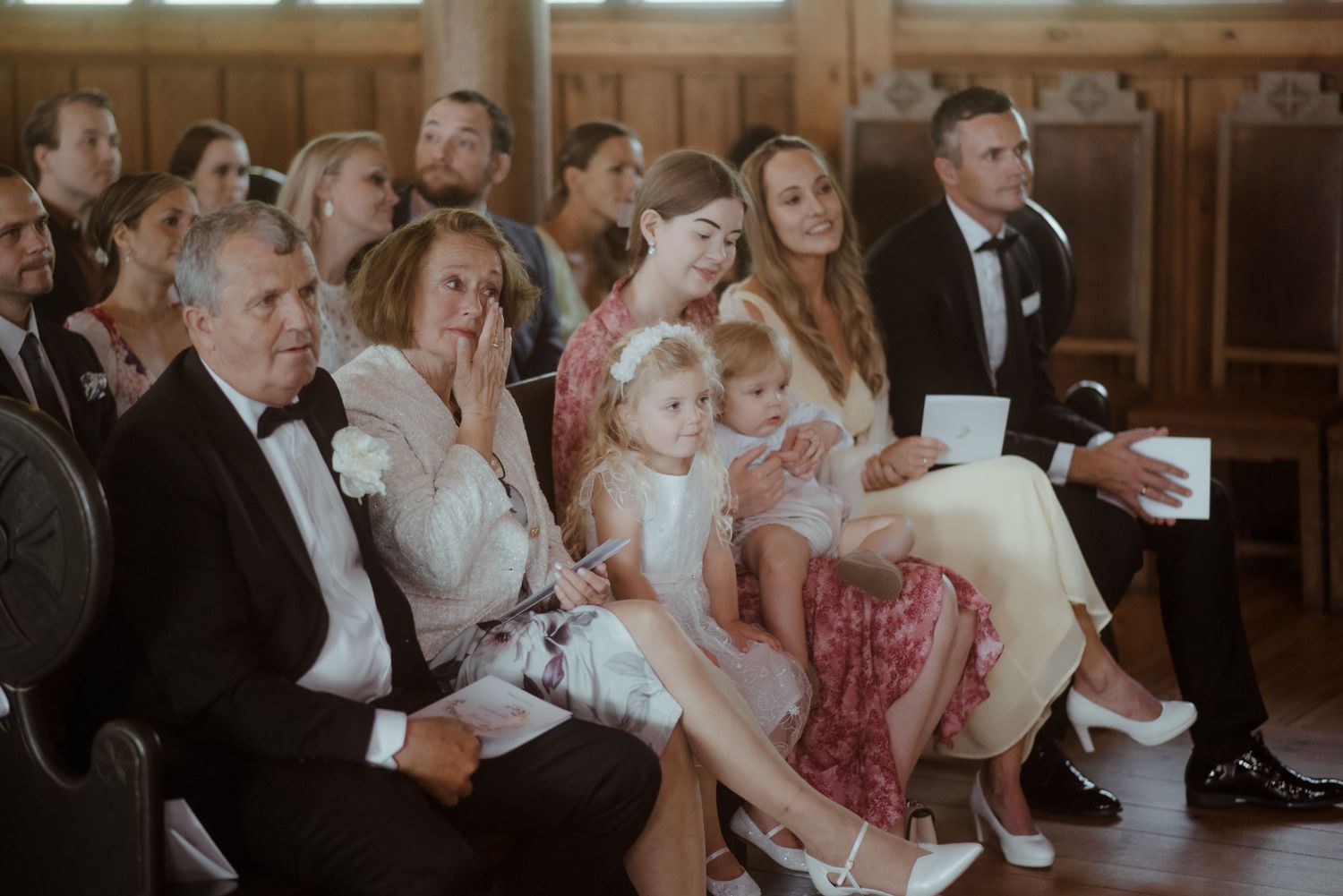 Wedding guests and children seated in wooden church pews during ceremony.