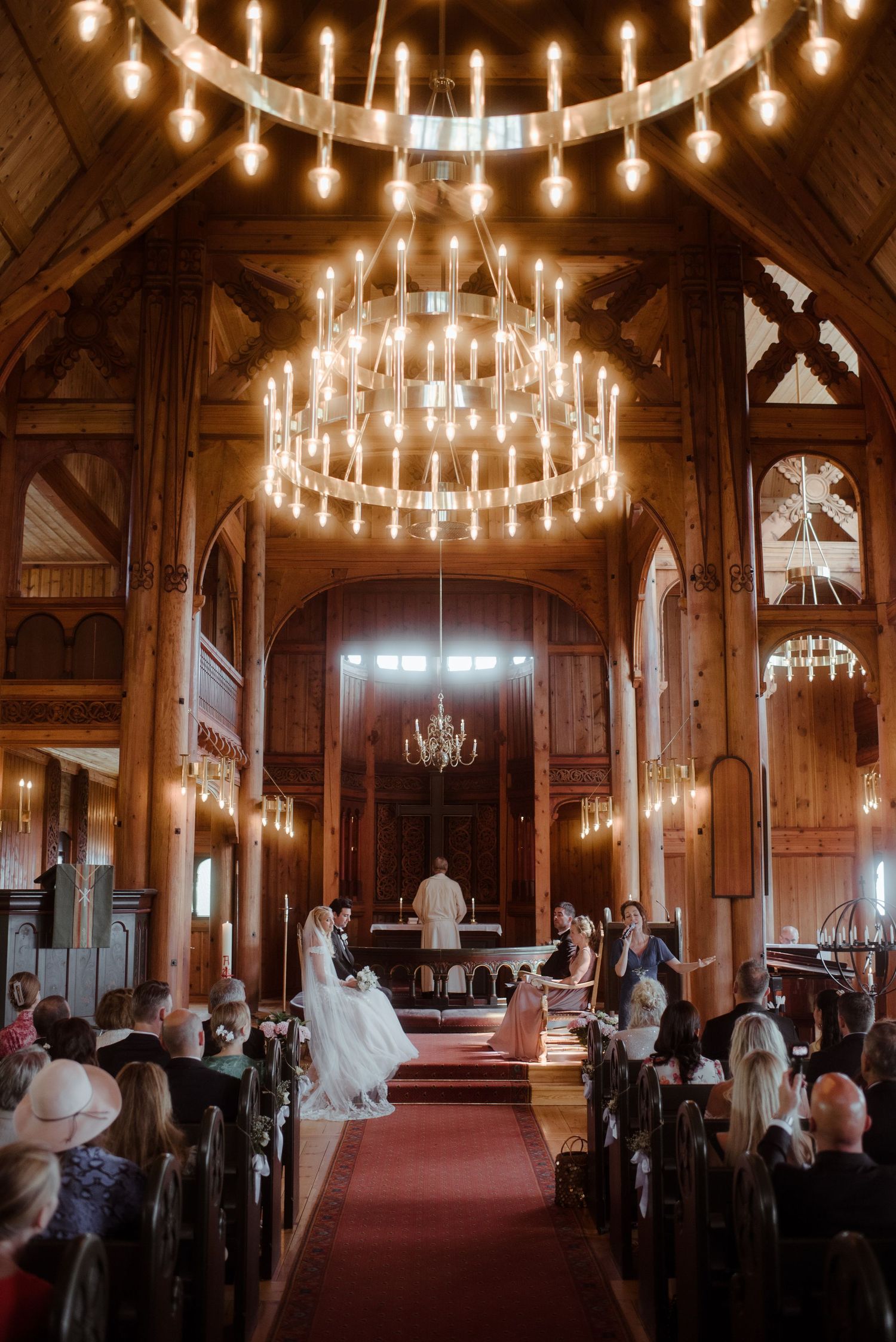 Wedding ceremony in a stunning chapel with multiple circular chandeliers and wooden beams.