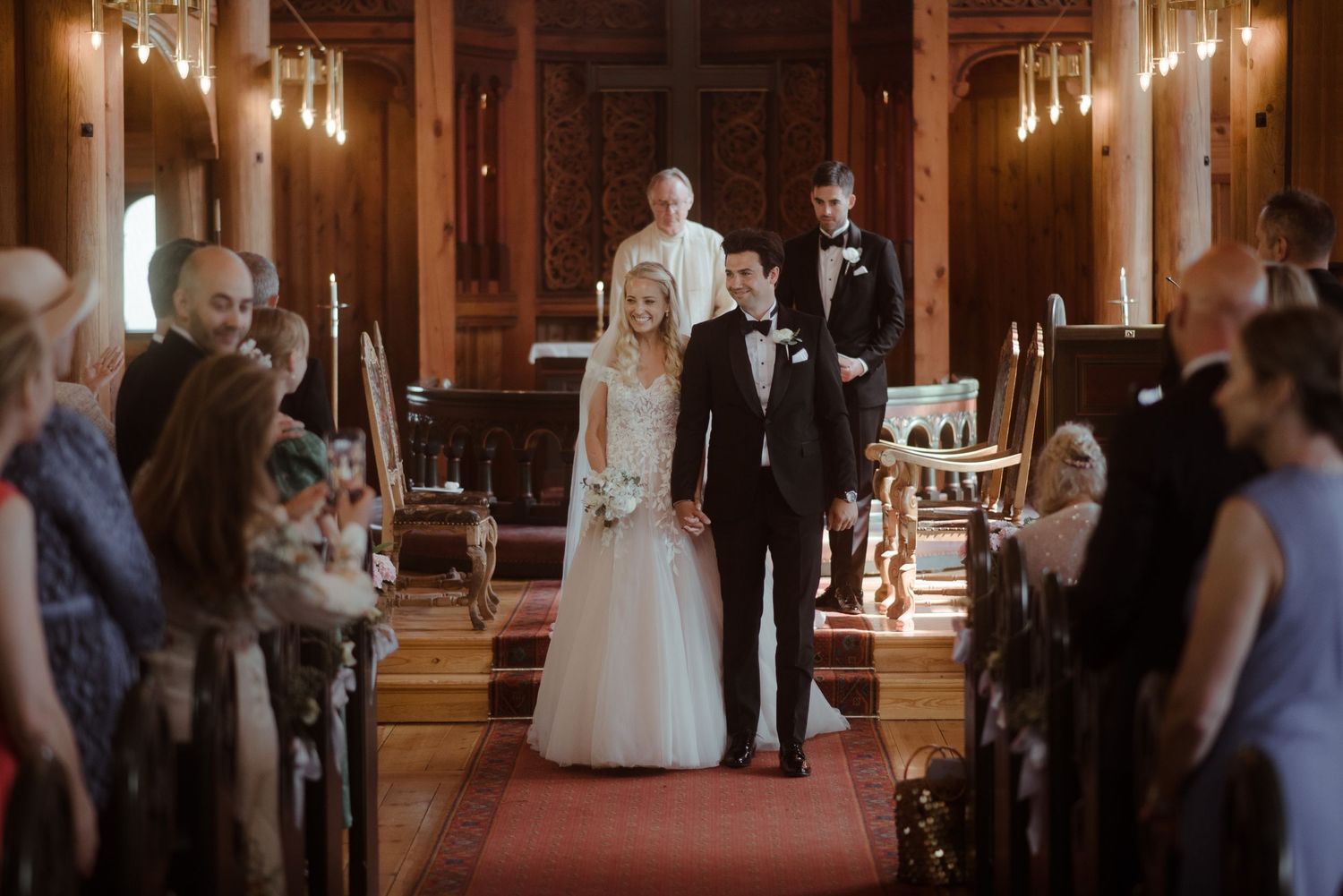A wedding procession walks down a red-carpeted church aisle lined with glowing candles and guests.