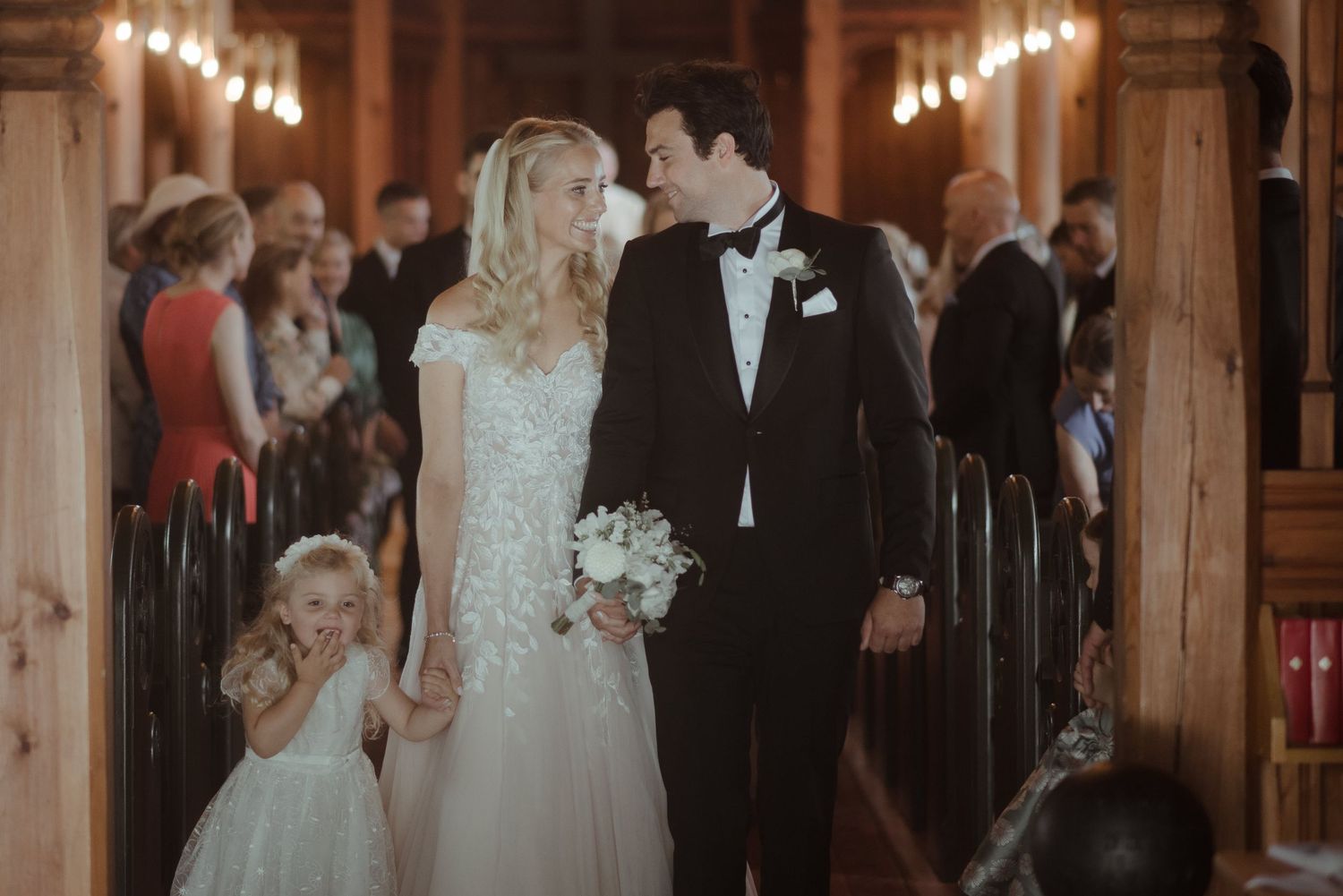 A wedding processional in a warmly lit church with guests looking on from wooden pews.
