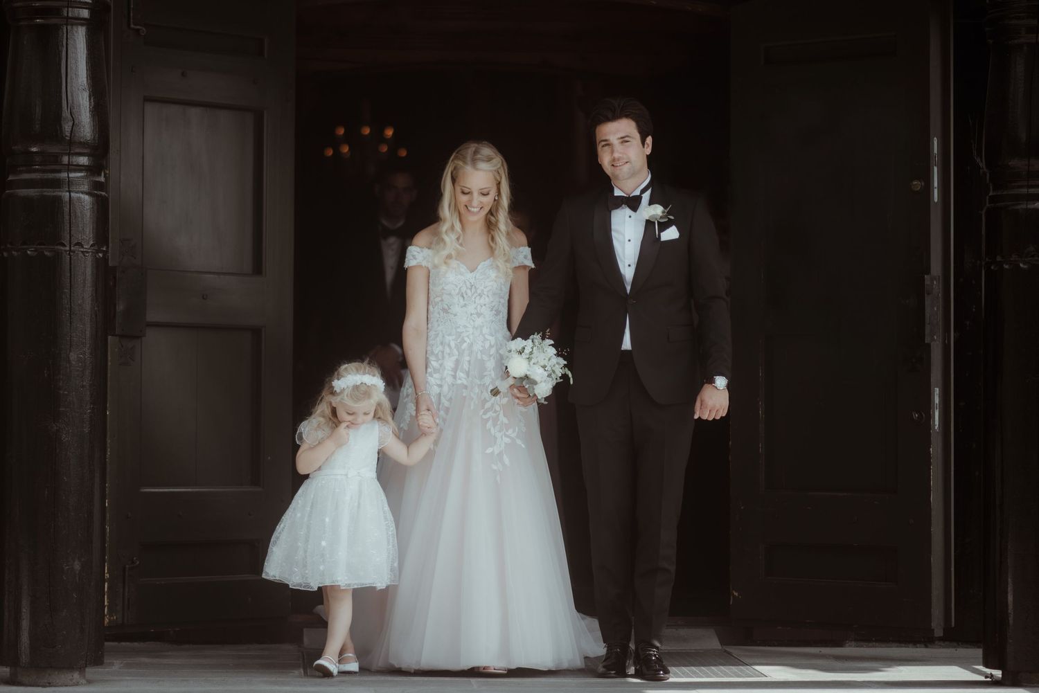 Elegant wedding portrait showing couple and young flowergirl in formal attire standing in church doorway.