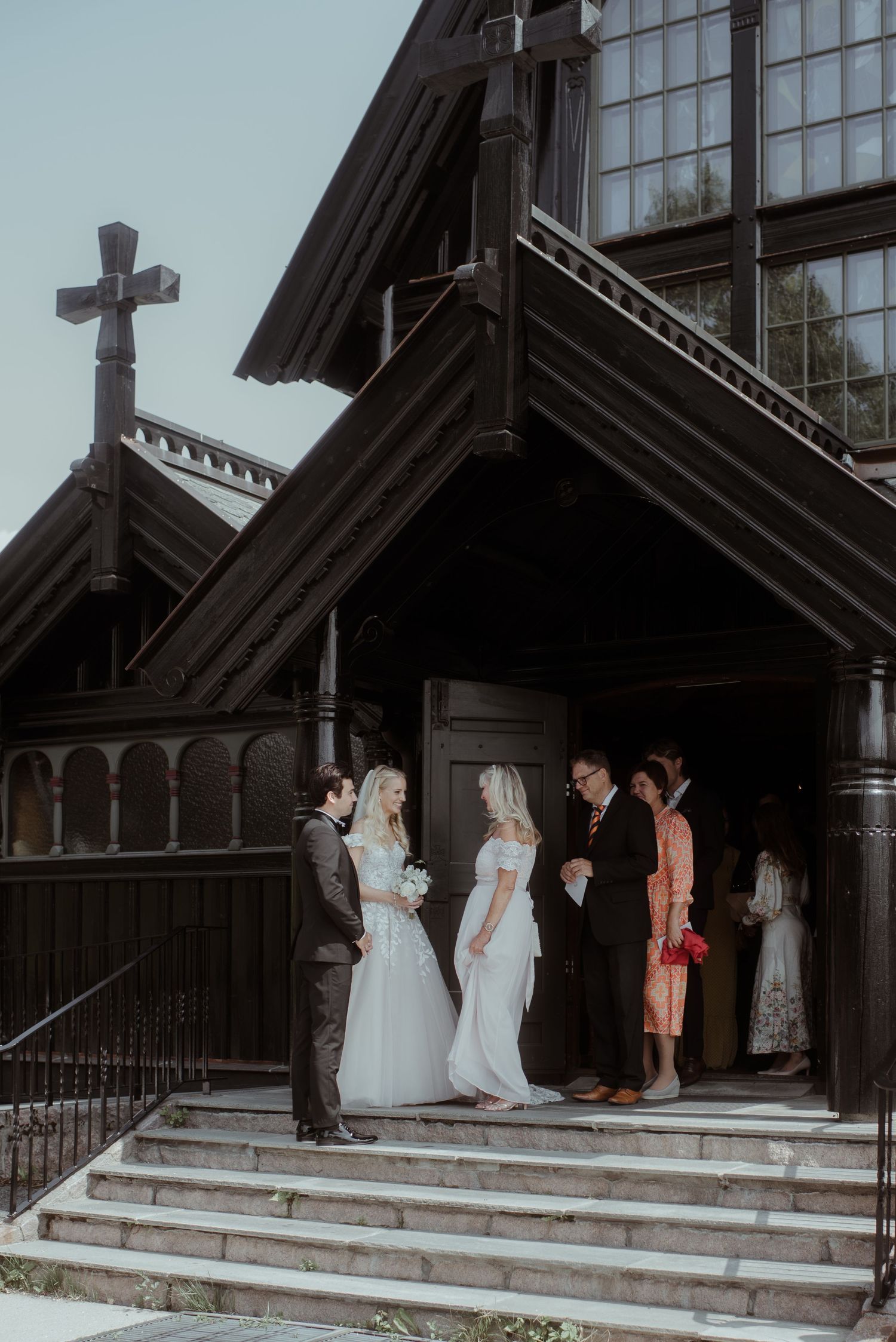 Wedding party gathered on steps of wooden church with distinctive cross on roof.