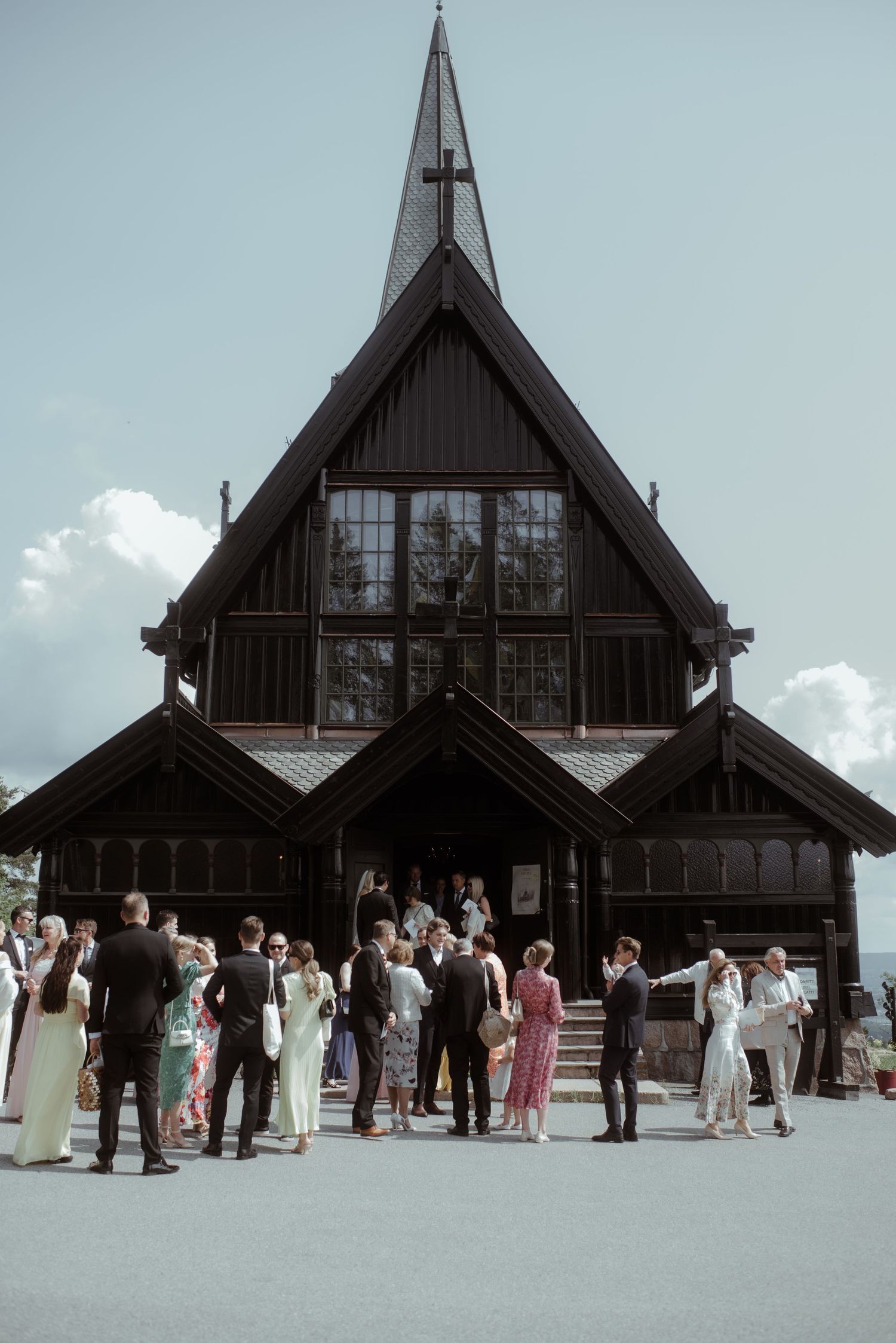 Large group of wedding guests gathered outside rustic wooden church with tall spire.