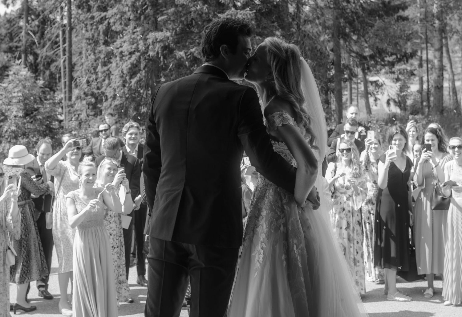 Newlyweds share a kiss surrounded by wedding guests in an outdoor forest setting.