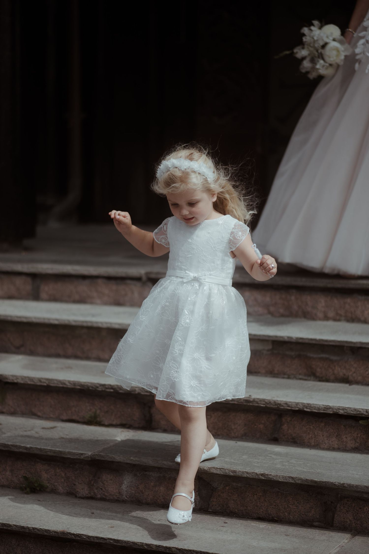 A young flower girl in a white dress dances on stone steps.