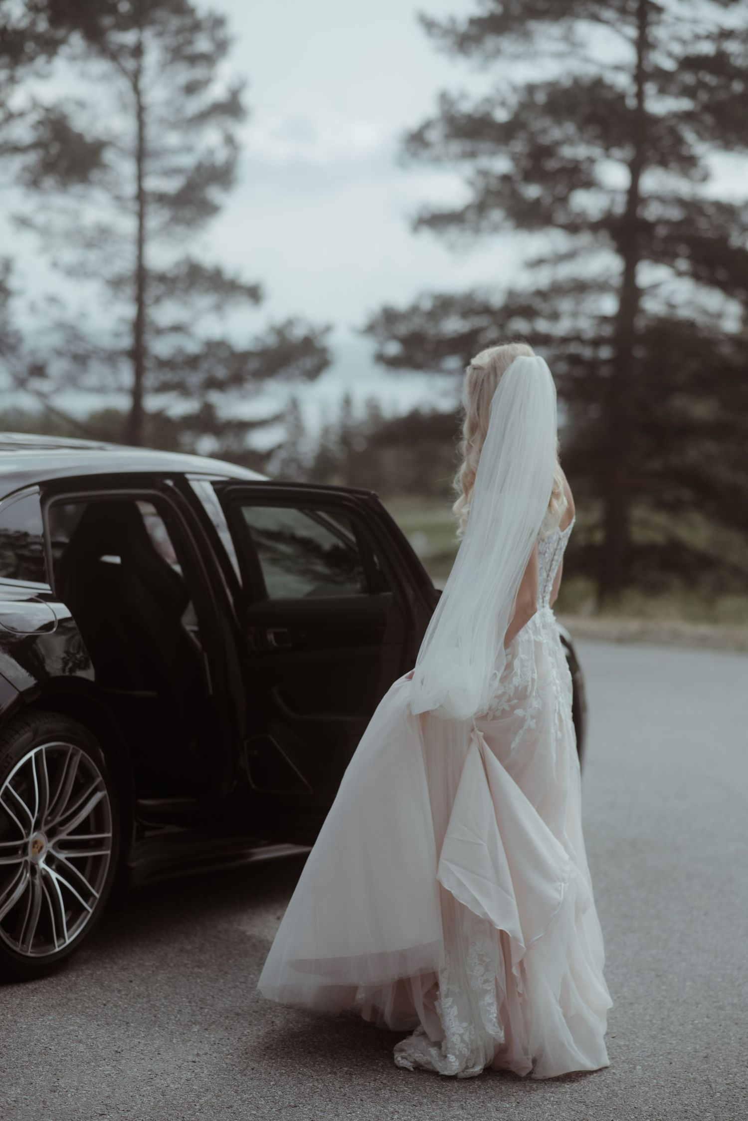 A bride in a flowing white dress stands beside a luxury car on a misty road.