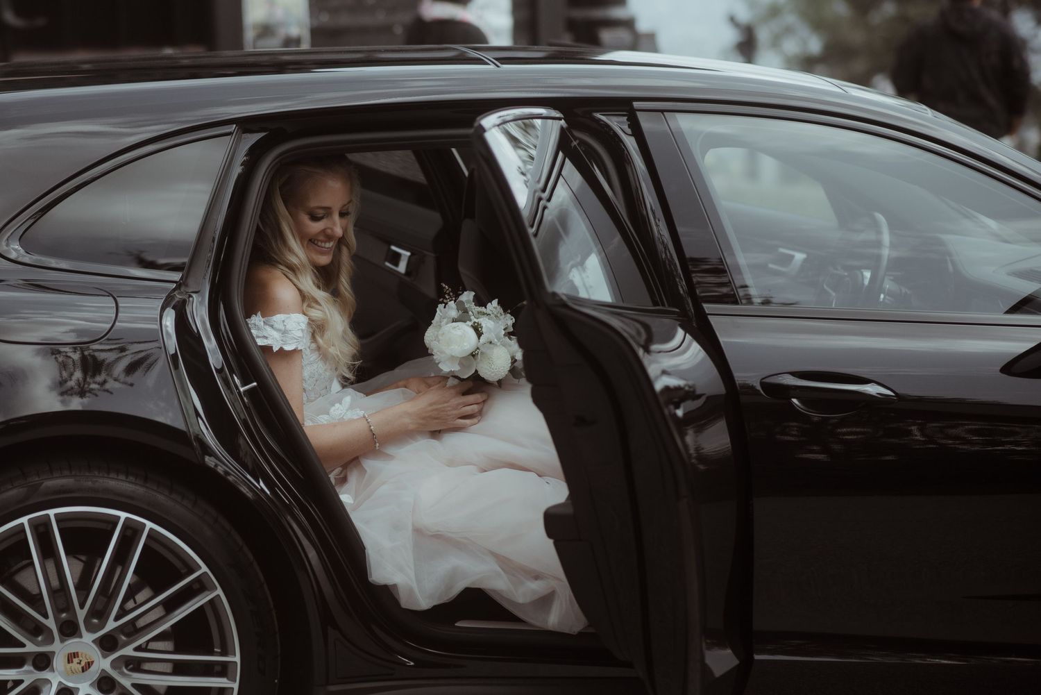 A bride in a white dress sits in the backseat of a black luxury car holding a bouquet.