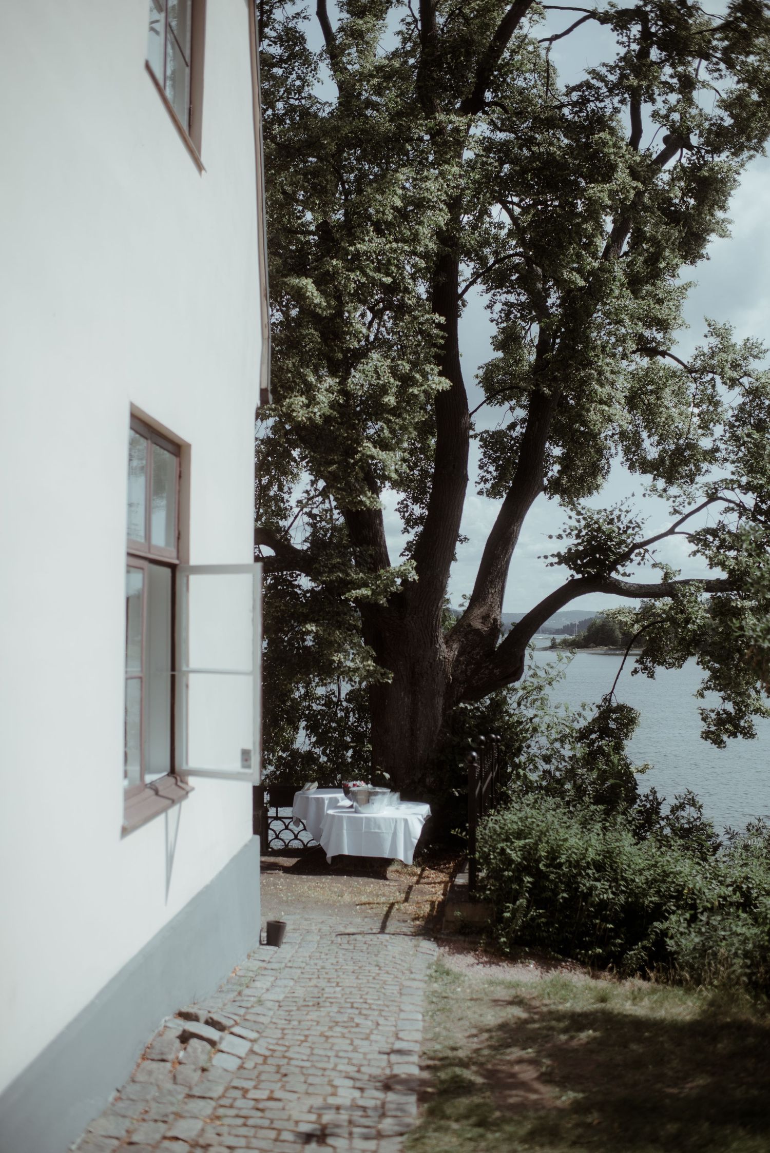 White building exterior with large tree and stone path leading to waterfront view.