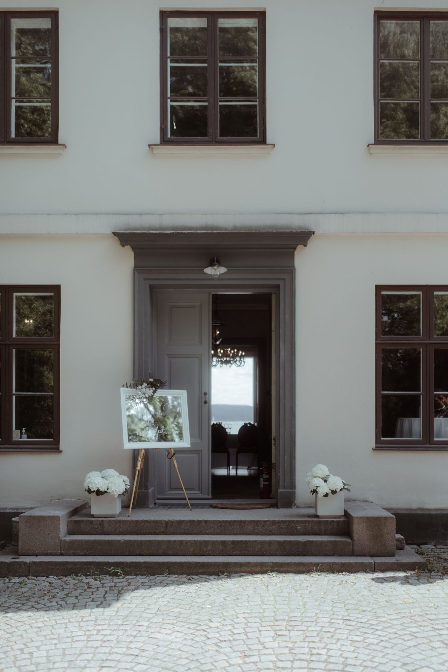 Classic white building entrance with grey door, windows, and potted flowers on steps.